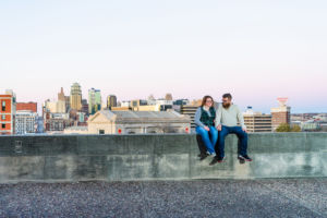 Dave and Bethany Strieff sit atop a stone wall with Kansas City skyline in the backdrop version 1